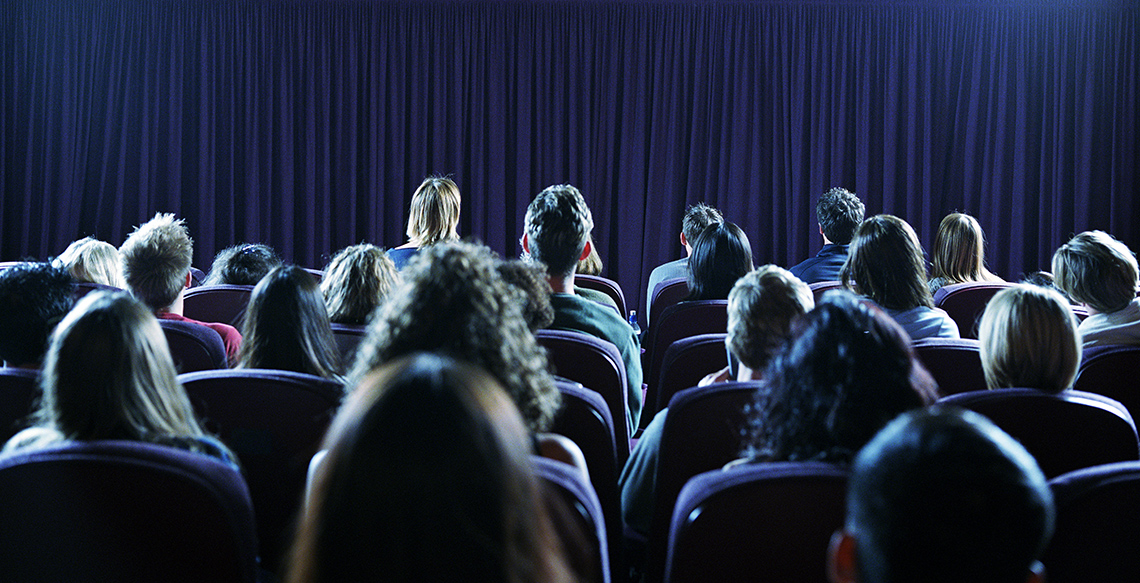 A crowd of people sit indoors in a theatre, in rows, facing away from the camera and towards a stage that is covered by a velvet curtain.