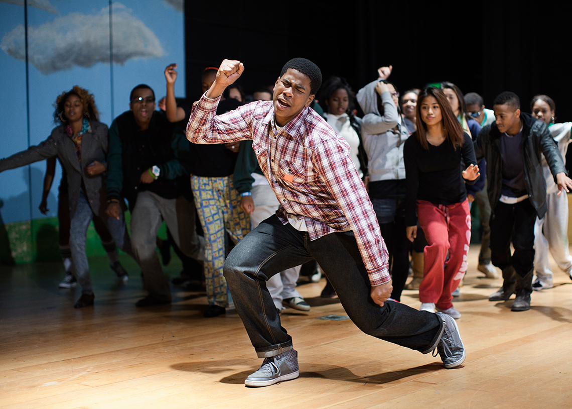 A dancer, their right arm raised, left arm lowered, and bending on one knee, leads a crowd of dancers forward in an indoor dance rehearsal.
