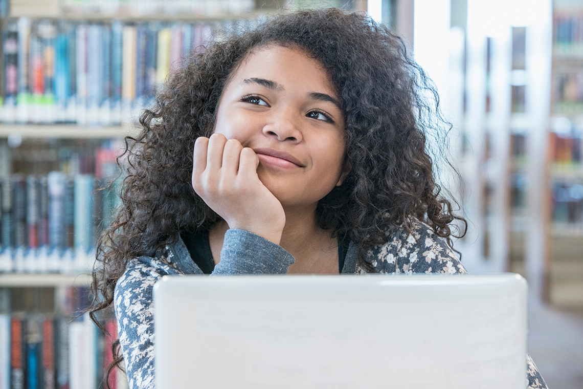 A student, their chin on their hand, sits looking up indoors in a library, rows of bookshelves behind them, a laptop open in front of them.