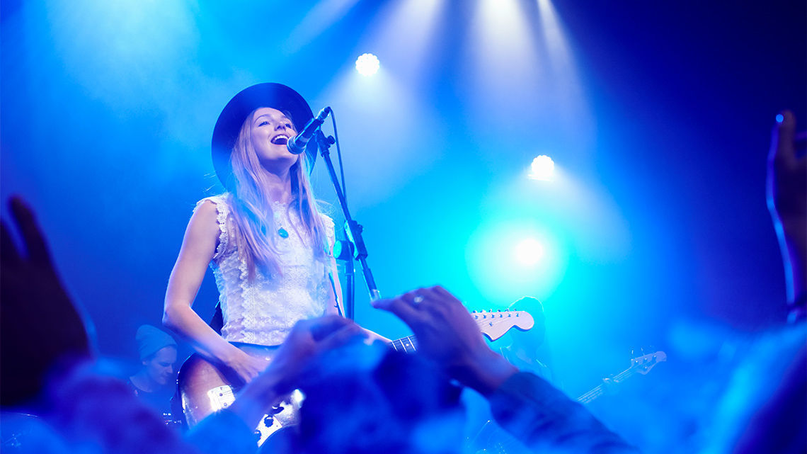 A singer in a wide-brimmed hat stands on stage indoors, with lights overhead, sings into a microphone, playing guitar in front of a crowd.