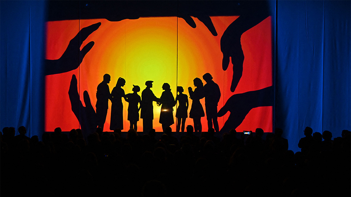 Eight actors stand behind a curtain on a backlit stage so that they are silhouettes. Silhouettes of hands form a circle around the actors.