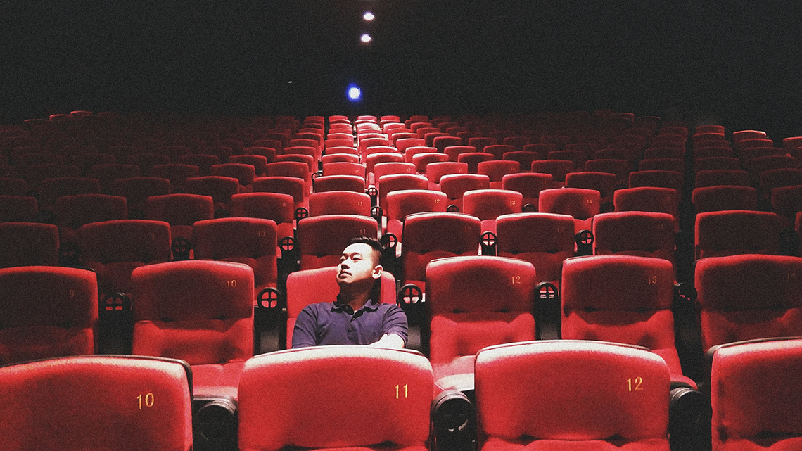 A young person sits alone in a dimly lit movie theatre, arms crossed in front of them, in a seat in the second row, looking up to the left.
