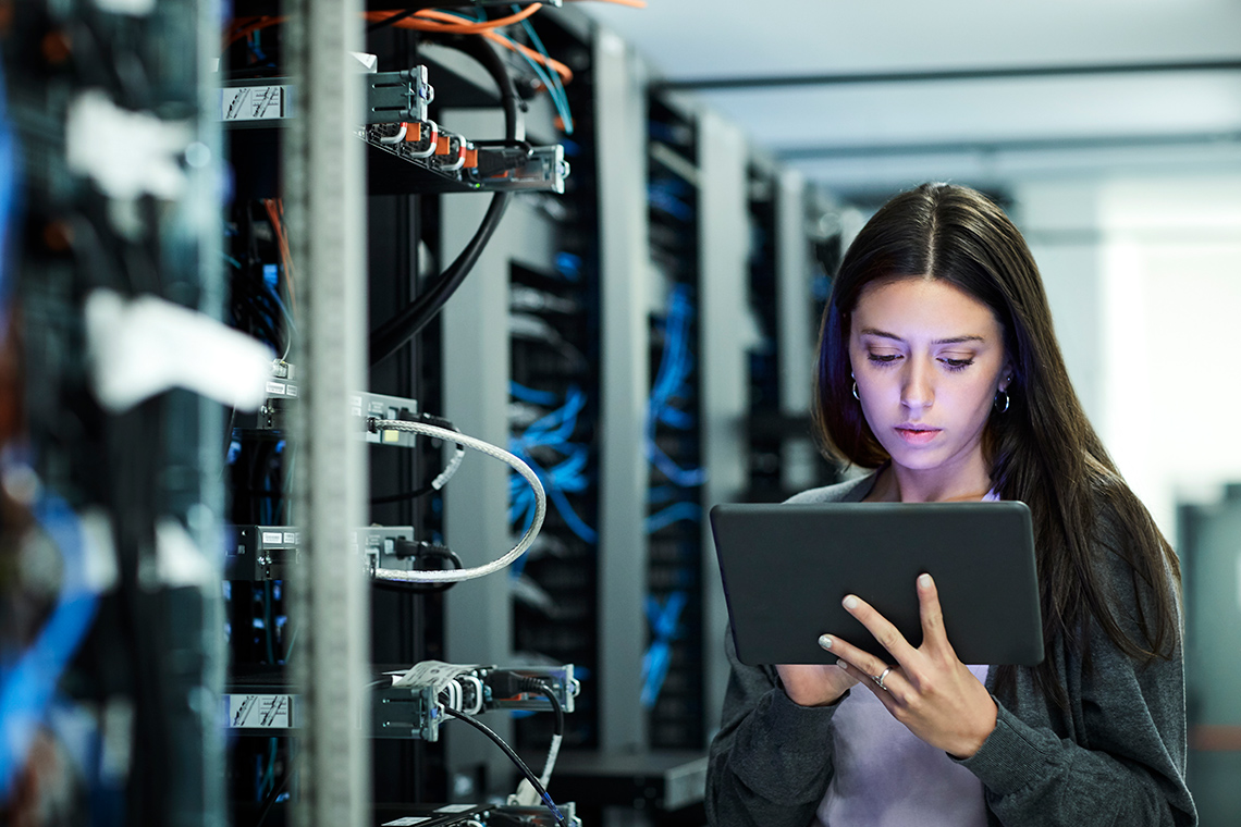 A young person stands indoors in a computer data centre, next to racks of computer servers with wires, looking at a tablet held in one hand.