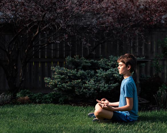 A student sits cross-legged on grass outdoors in a yard next to a fence, bushes and trees, back straight, eyes closed, hands open on lap.