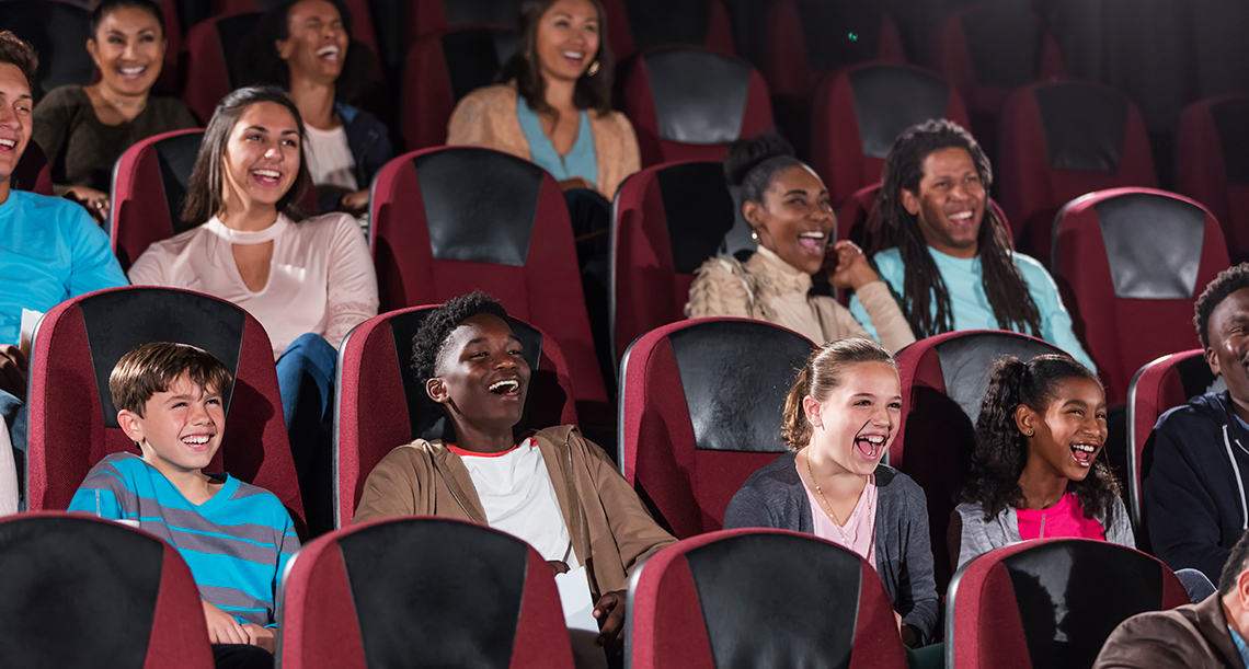 A group of young people sit in several rows of seats indoors in a theatre, facing the stage, laughing and enjoying the performance.