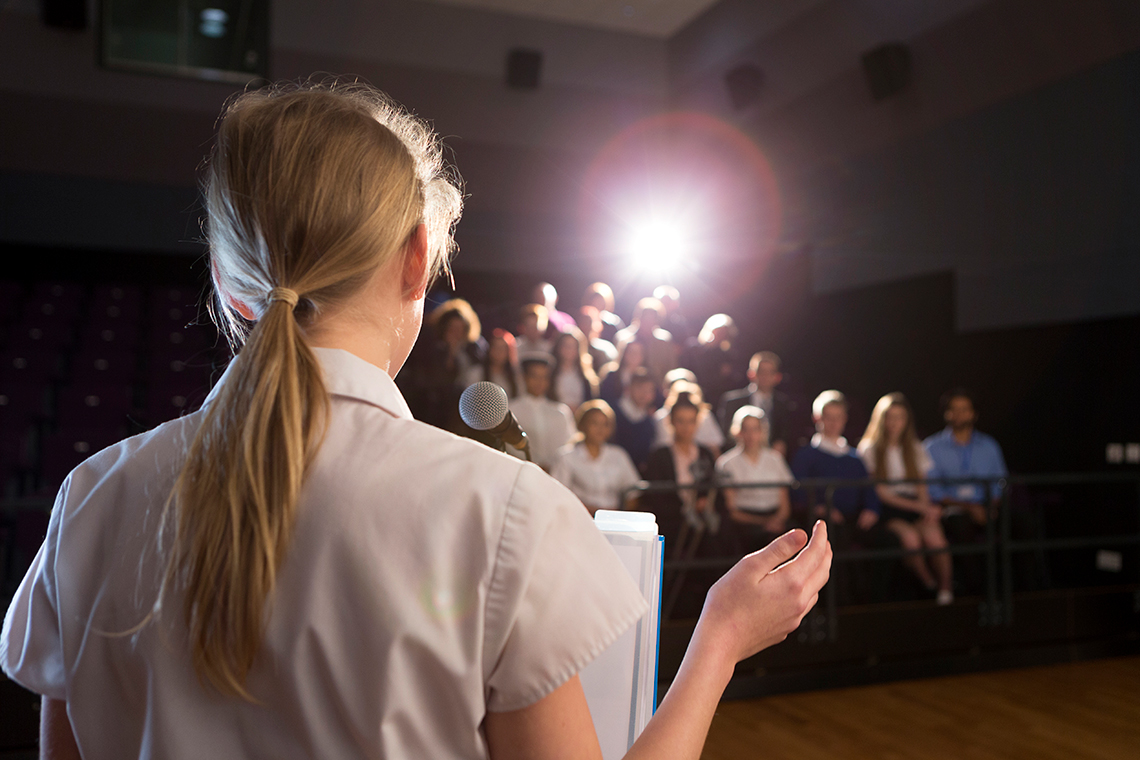 A young person stands on an indoor stage in a theatre, facing away from you towards an audience, talking into a microphone, making a pitch.