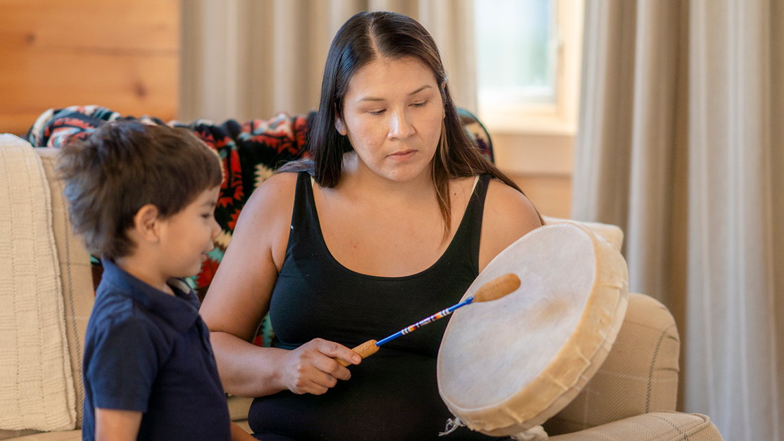 A young adult is playing a traditional drum for a child at home.