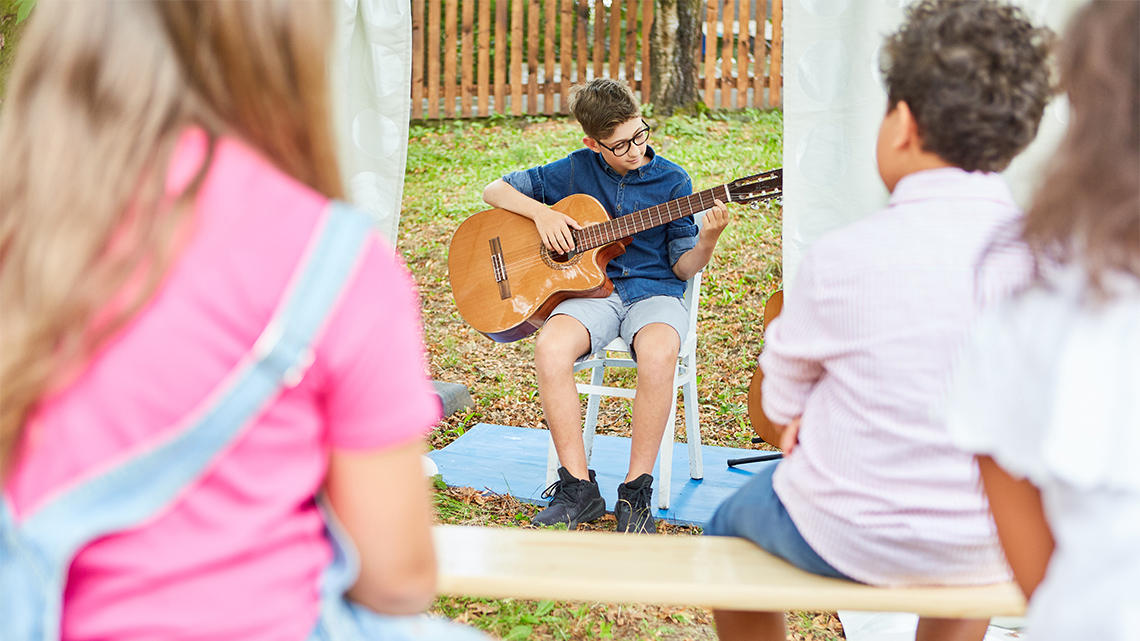 Kids sitting and watching another child play the acoustic guitar.