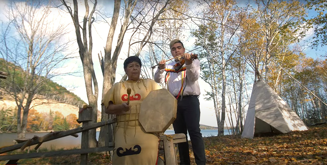 Morgan Toney plays the fiddle in the woods. Another musician plays a traditional Mi’kmaq drum. 