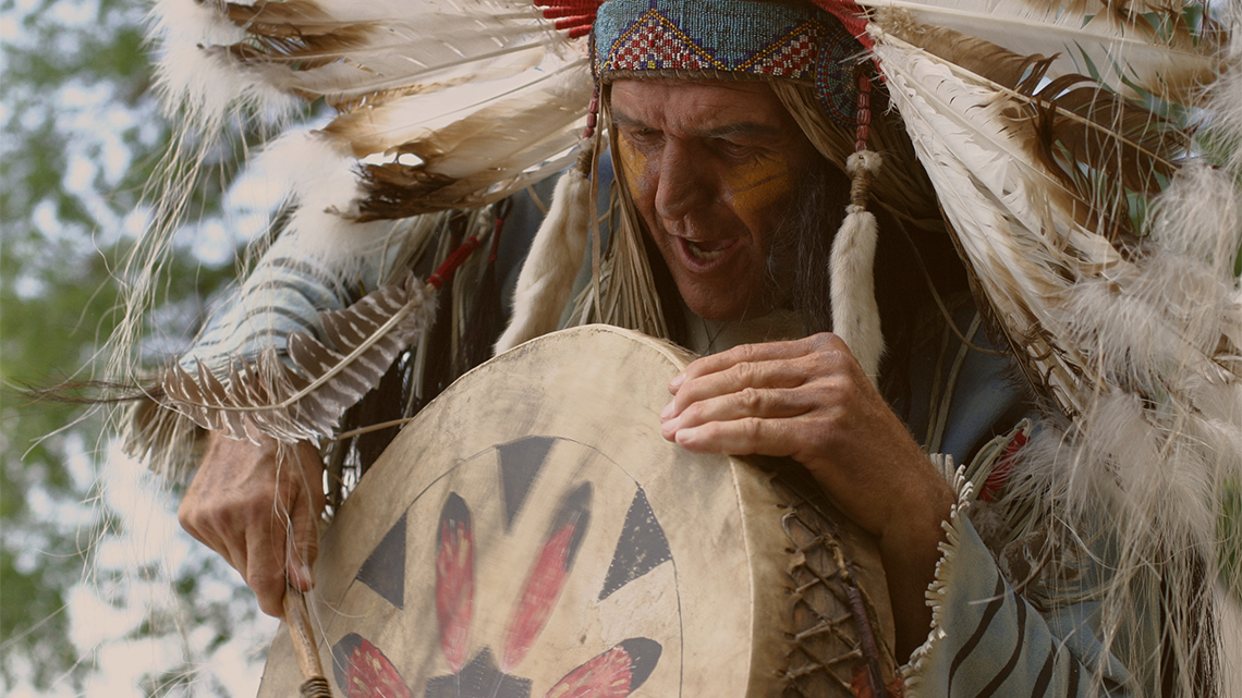 An Indigenous in buckskin and feather headdress plays a drum as he chants.