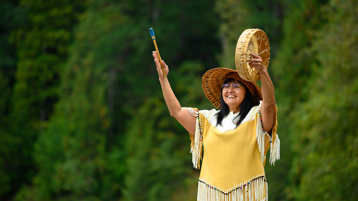 An Indigenous person is playing a hand drum with a mallet.