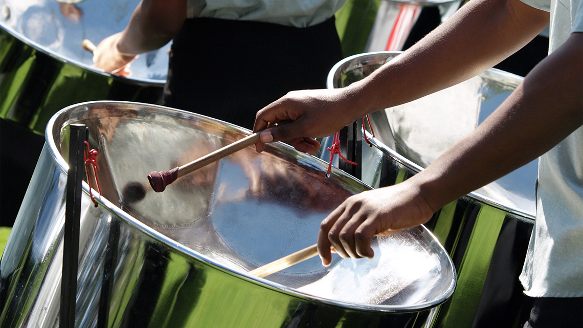 A person playing a curved steel drum with two mallets outside with other musicians.