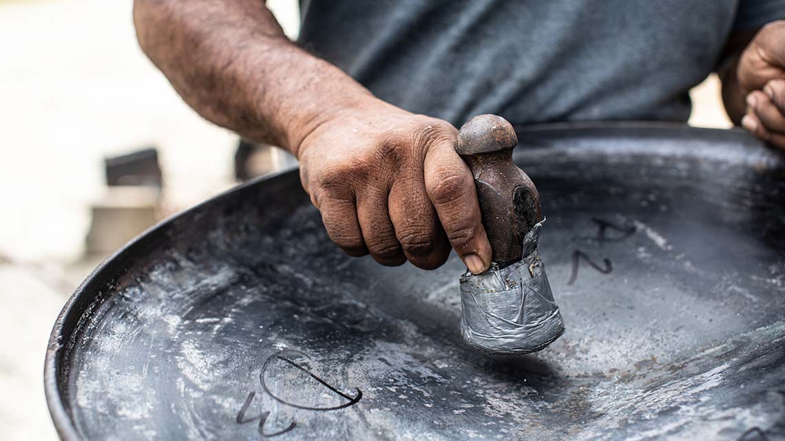 A person hammering a steel drum to create dents in the bottom surface.