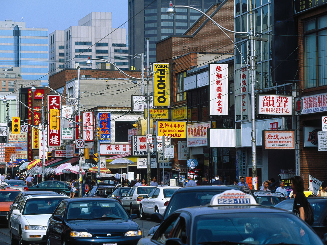 A busy city street lined with many businesses. People are walking on the sidewalk and there are several cars on the road.
