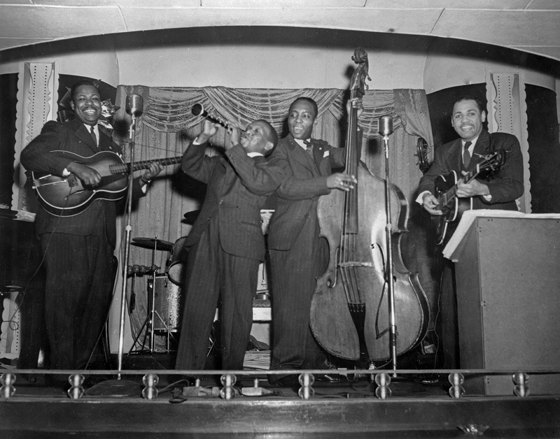 Four Black musicians on a stage playing instruments such as guitar, clarinet, and upright bass.