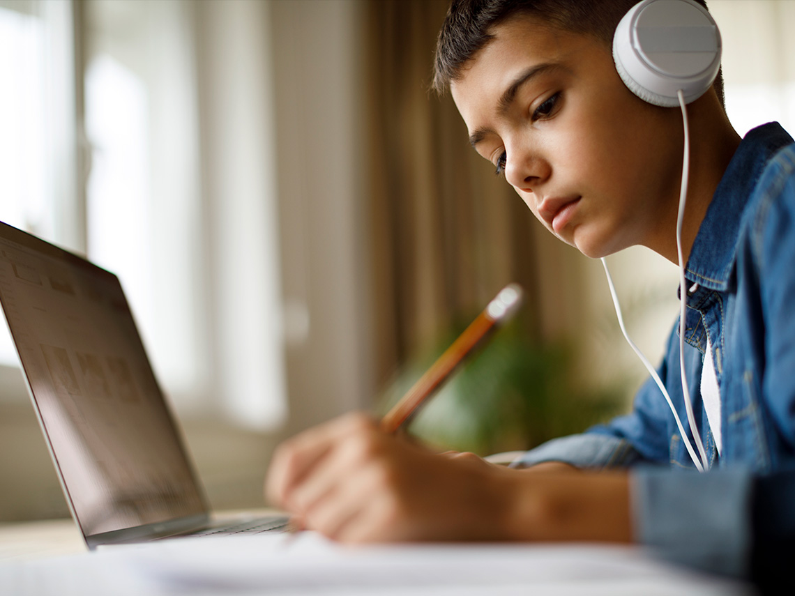 Student with headphones on and a computer in front of them writing in a notebook annotating music.