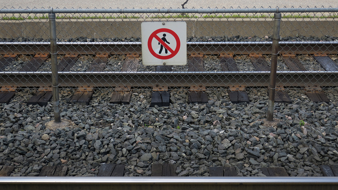 Sign on the fence beside a train track is a white background with a red circle and line through a person walking.