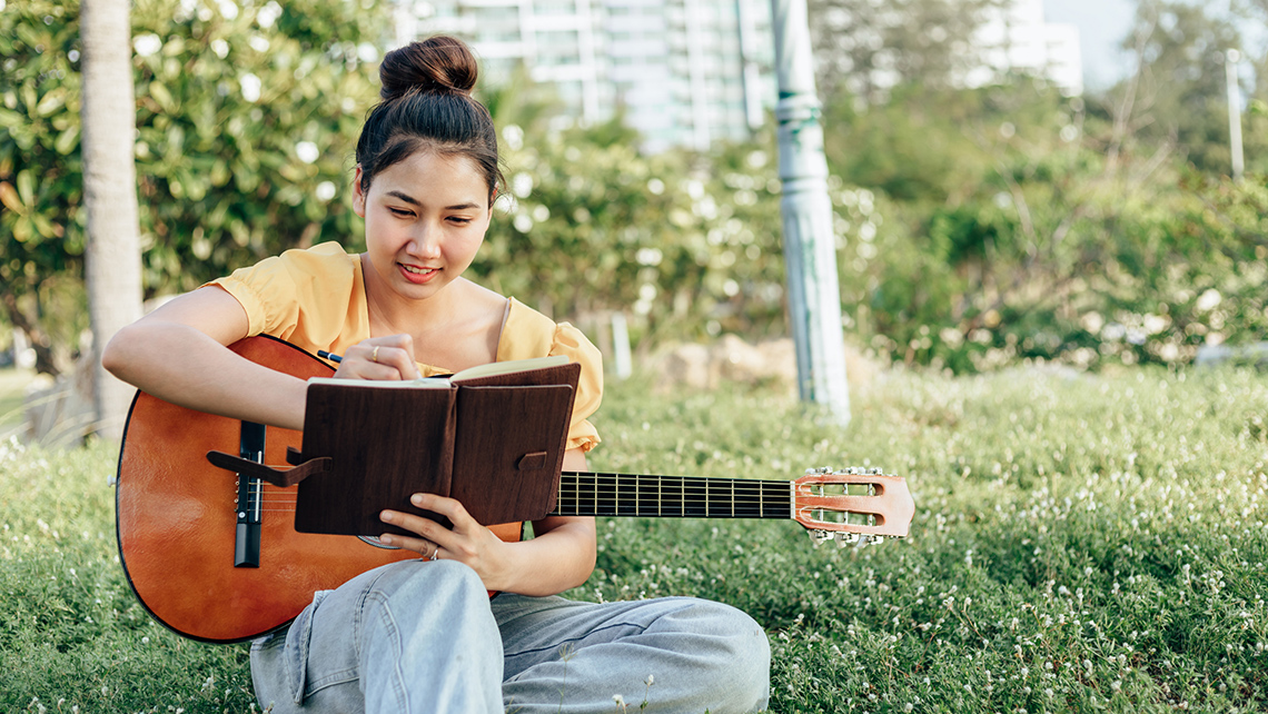 There is a young person outside in a park space holding a guitar in their lap and writing in a notebook. They are smiling.
