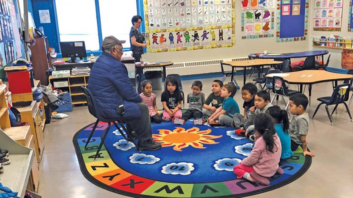 Students in their classroom sitting in a circle on the carpet facing an elder seated in a chair.