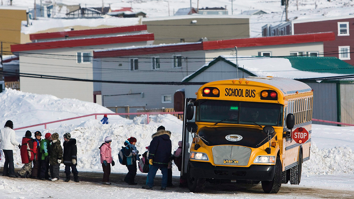 Students lining up on a winter morning to get onto the school bus. There are houses and snow.