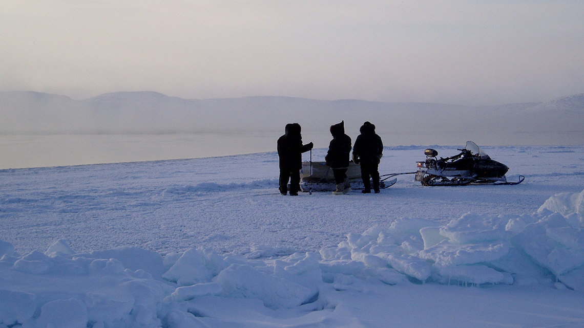 3 people in the snow standing in front of a sled with cargo that is attached to a snow mobile.