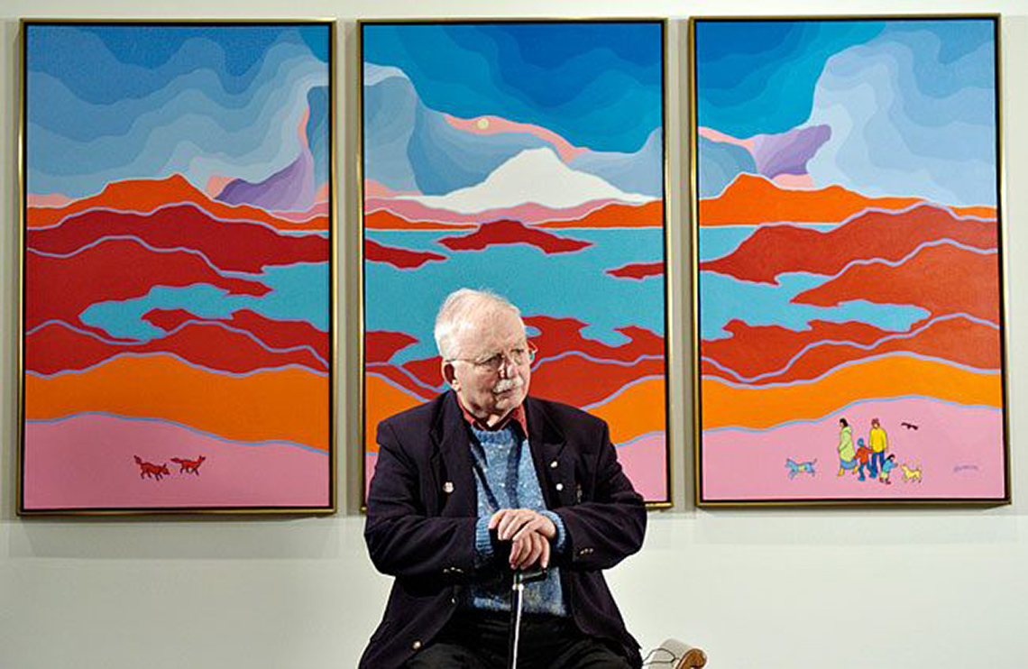Ted Harrison sitting in front of his paintings in a gallery