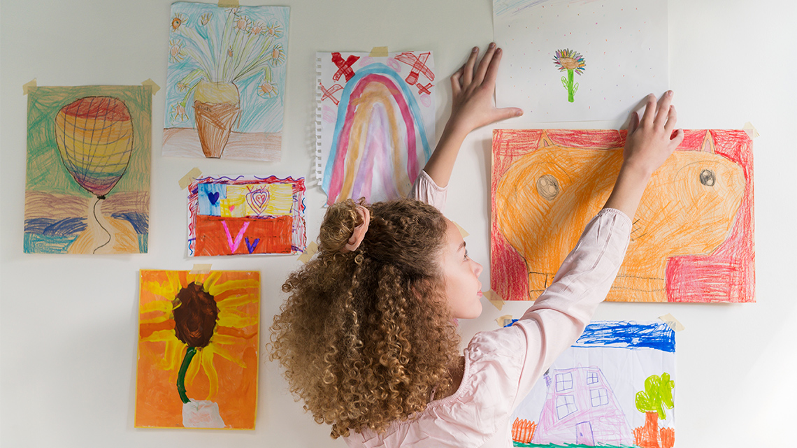 A child hanging a drawing of a flower on the wall amongst other pieces of artwork they may have created. 
