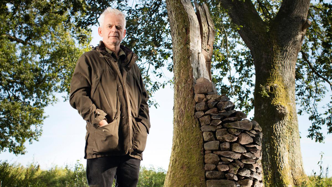 A photo Andy Goldsworthy standing outside in front of trees. He is wearing a jacket and his hands are in his pockets.
