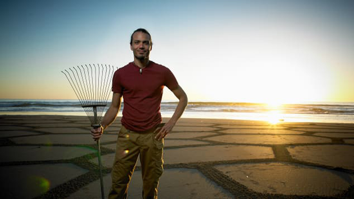 Photo of Andres Amador holding a rake in front of one of his art pieces.