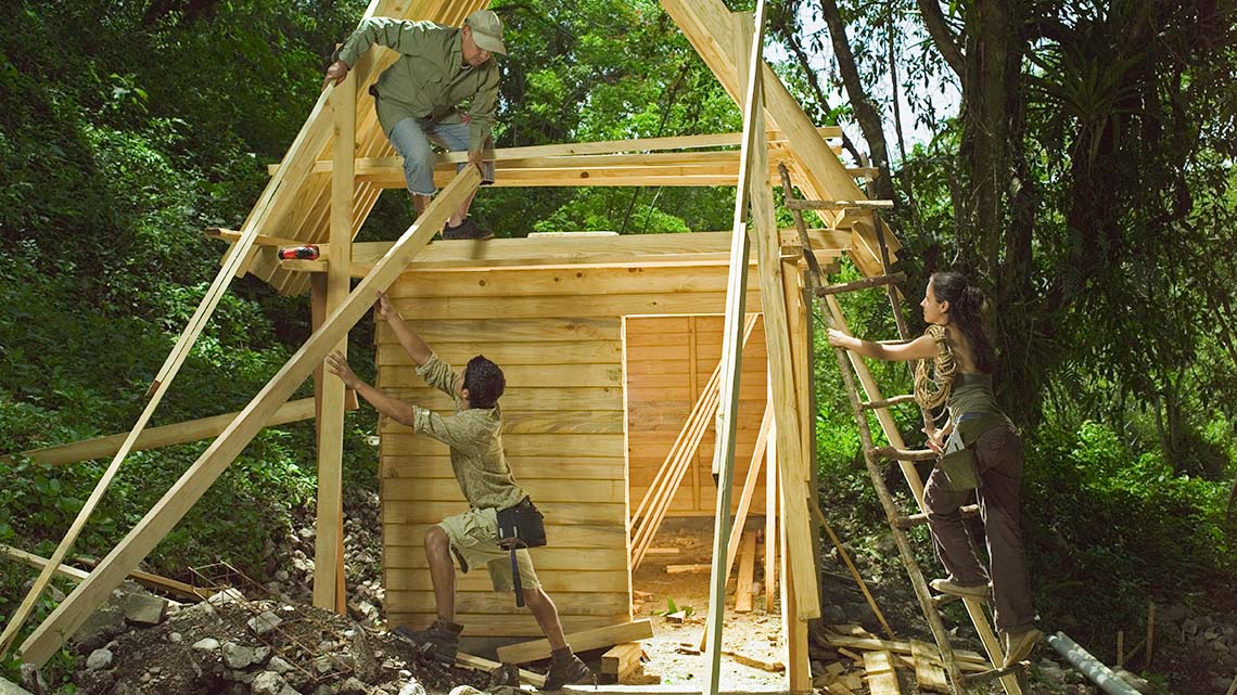 Three people constructing a small, eco-friendly house out of wood in a forest.