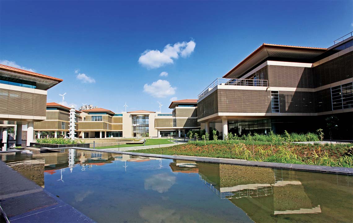 A group of buildings surrounding a grass courtyard. There are mutliple wind turbines behind the buildings.