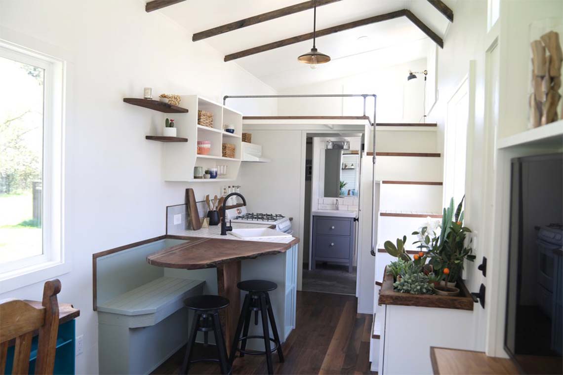 The inside of a tiny home with a kitchen, dining table, and a sleeping area built above a bathroom.
