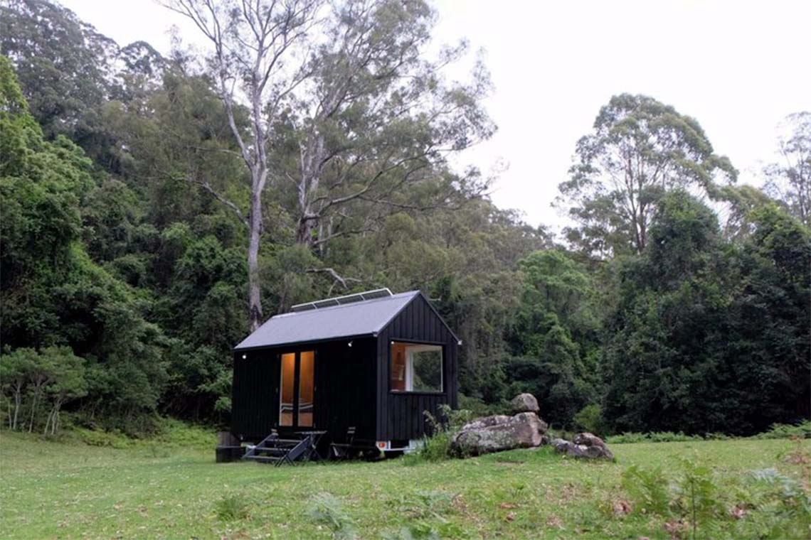 A tiny cabin in a field surrounded by trees with a solar panel on its roof.