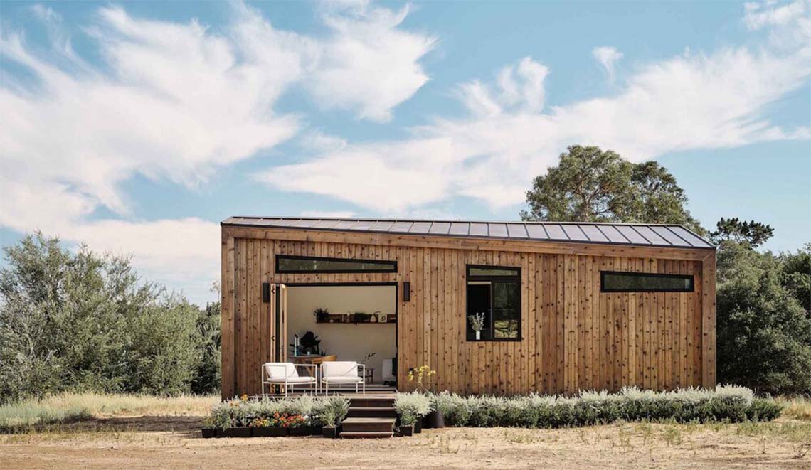 A tiny home in a desert surrounded by shrubs with two chairs on a porch.