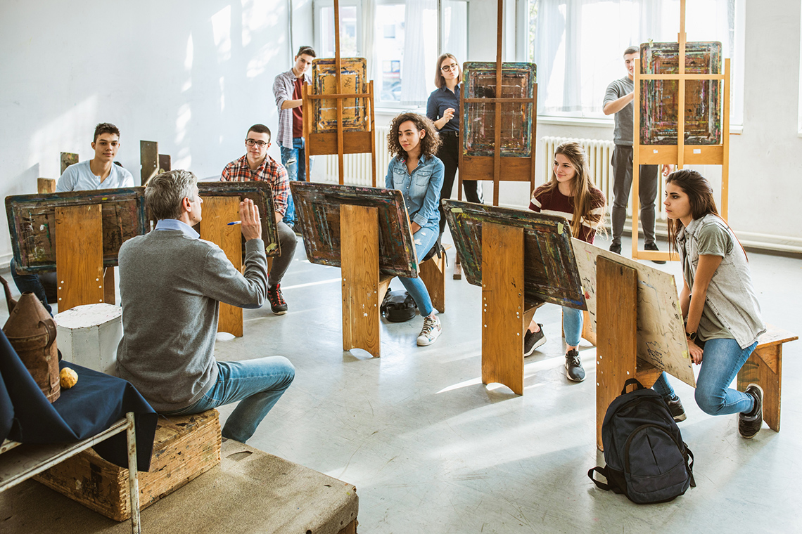 An adult instructing a group of youth sitting behind their own canvases on easels.