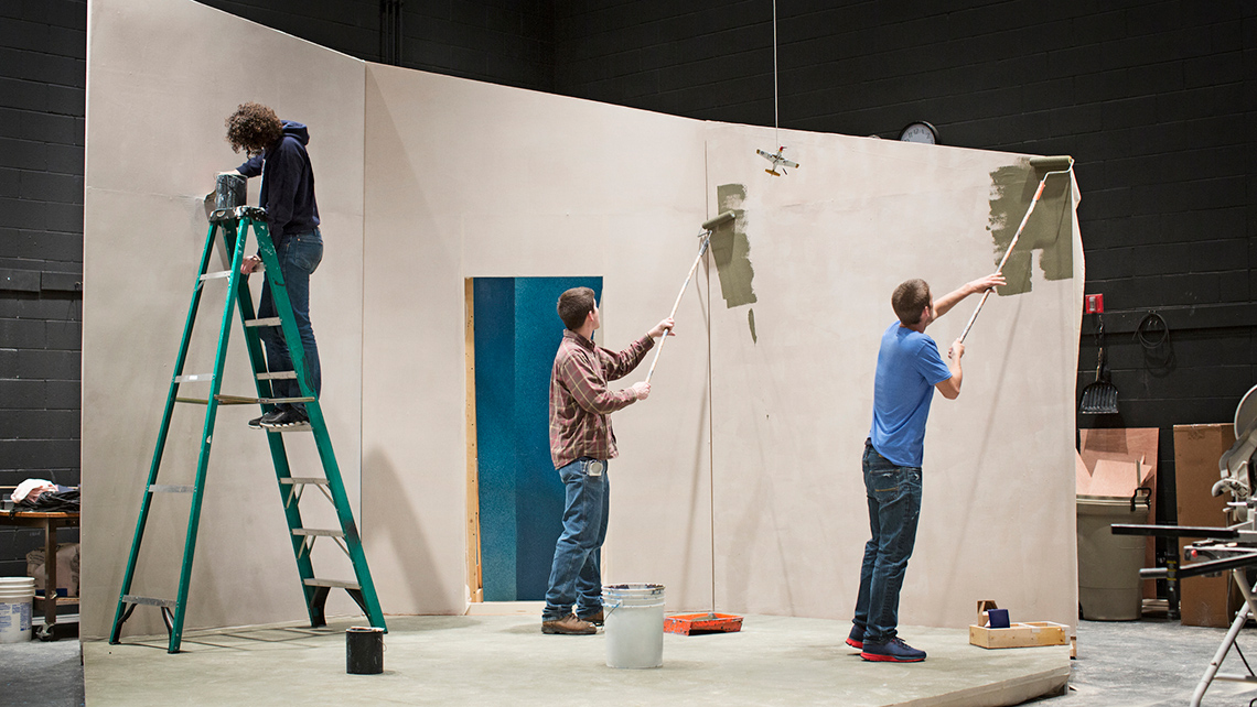 Three workers apply paint to the walls of a small set being built in a large studio space.