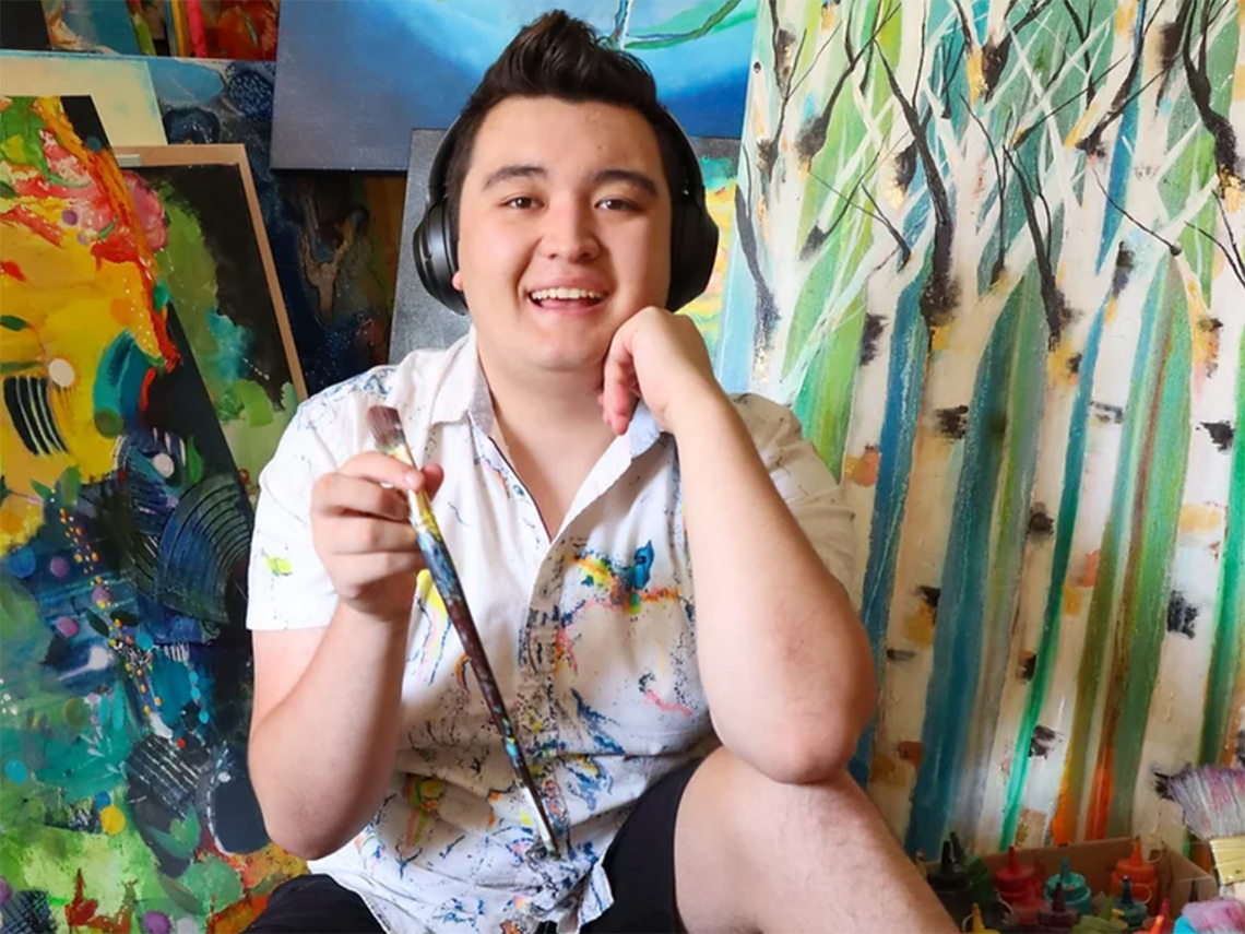 A smiling young man, wearing headphones and holding a paintbrush, against a backdrop of various artworks.