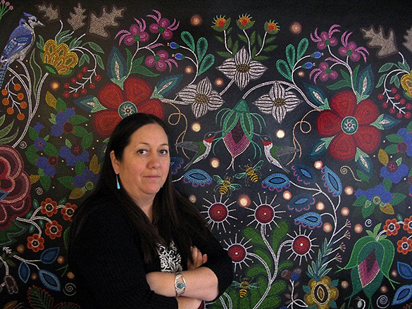 A portrait of Christi Belcourt in front of their artwork.
