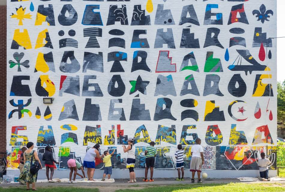 A large mural on the side of a building. The mural is being created by community members.