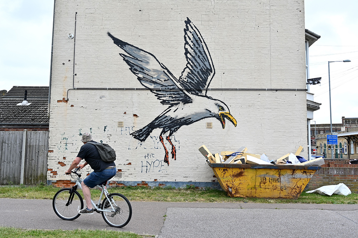 A spray-painted seagull on the side of a building appears to be landing on a real garbage dumpster.