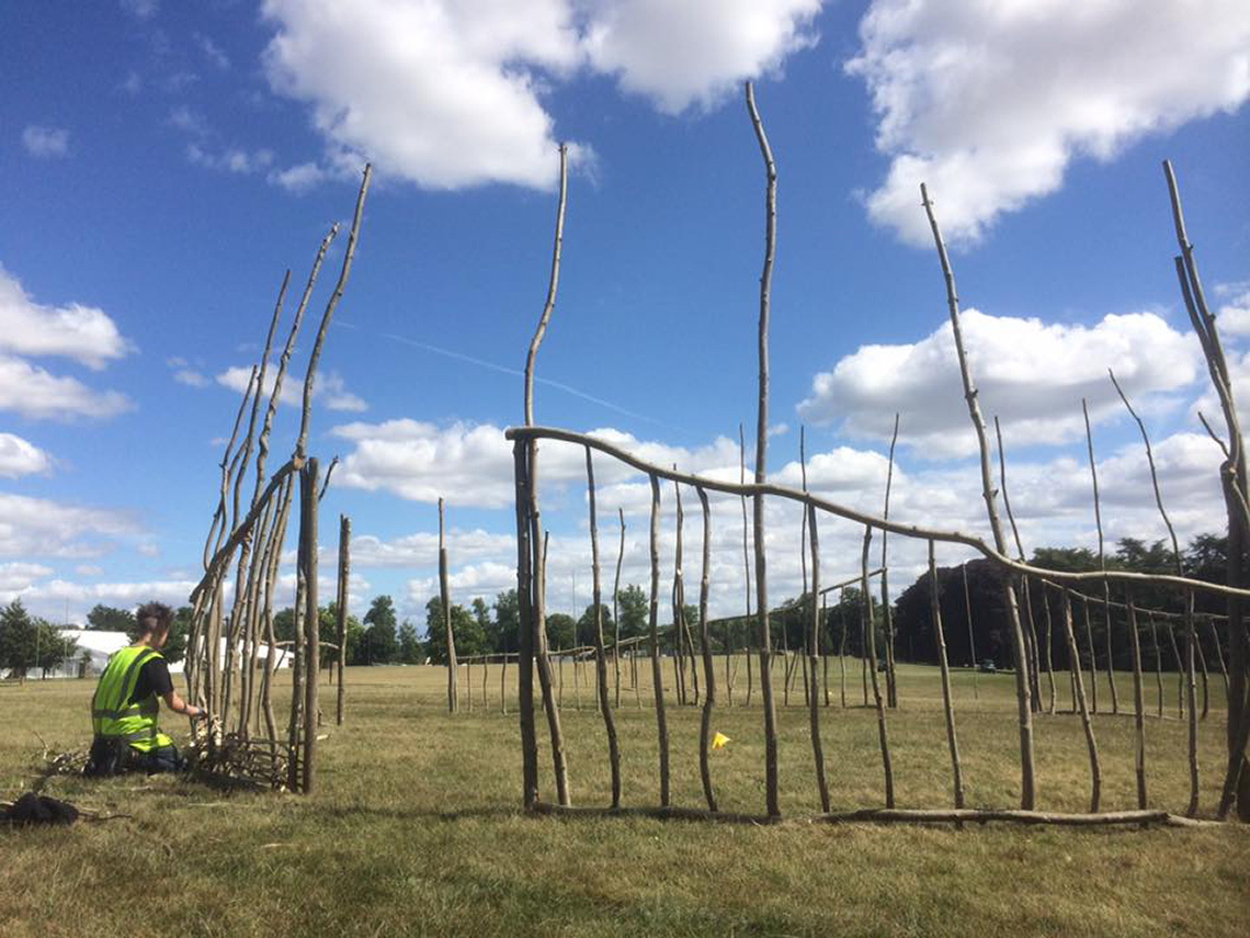 A sculpture of straight branches create a fence that covers the perimeter of an open grass space.