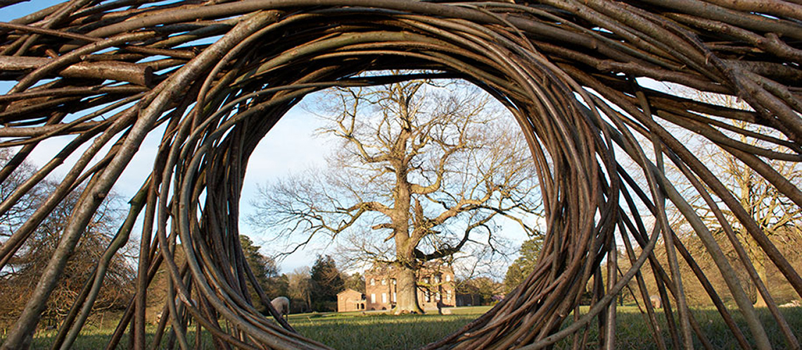 A circular sculpture made of branches with a round window. A tree is in the window’s background.