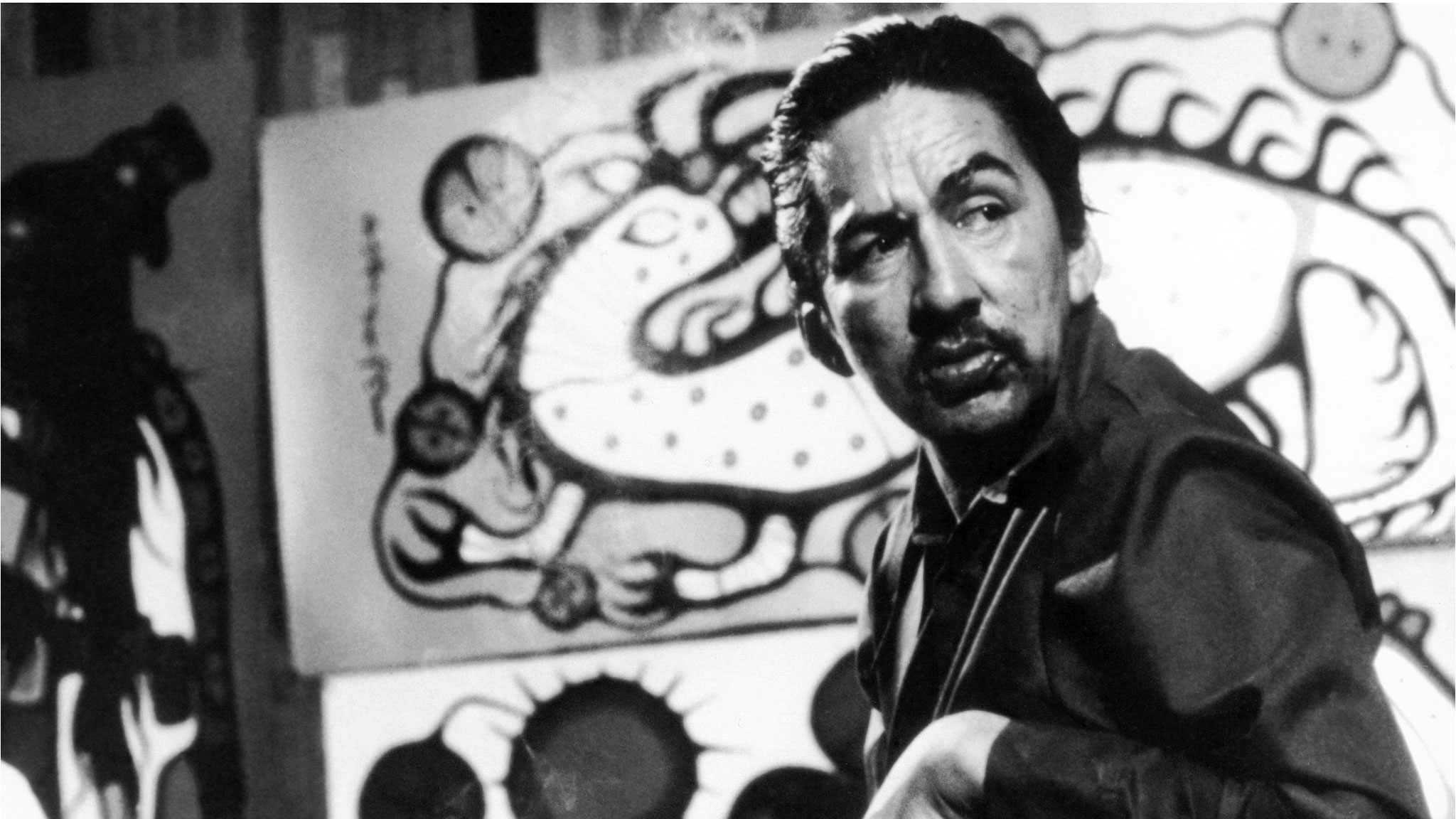 Norval Morrisseau sitting in front of paintings in his studio.