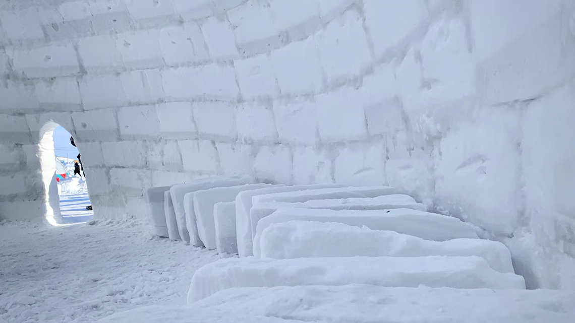Blocks of snow lined up inside of a large igloo. Sunlight shines through the doorway.