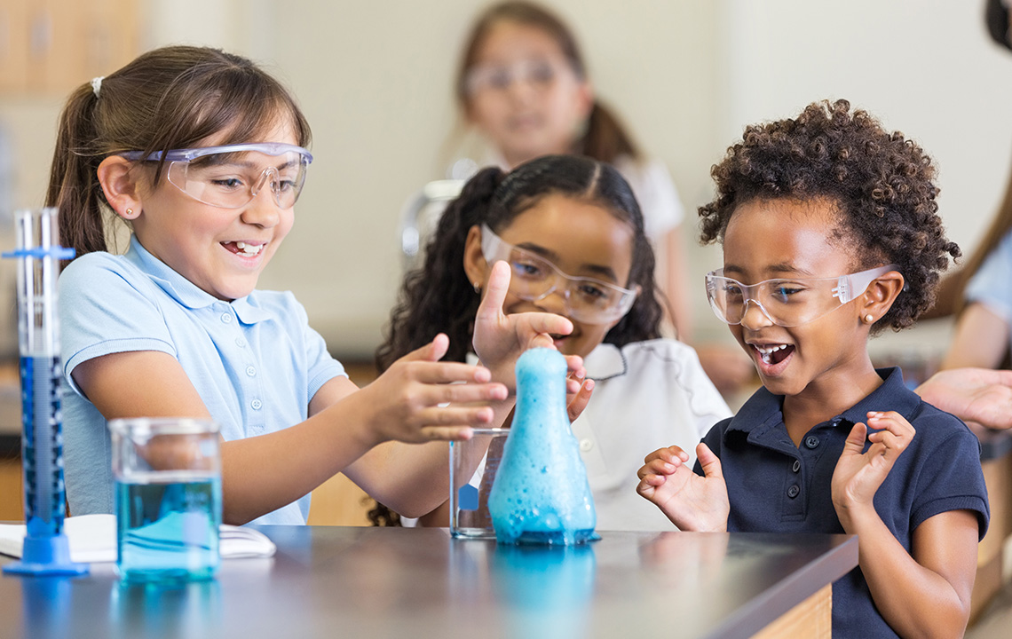 Three children conducting a science experiment in class. They are looking at a flask that has a foaming, blue liquid.