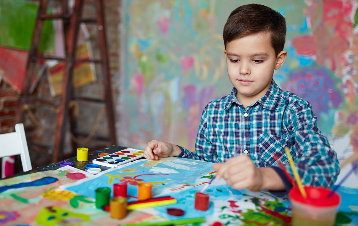 A student painting on a paper at a table. There are paints and brushes all around the table.