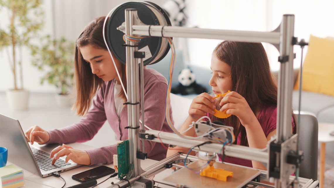 Two young students working together on a laptop by a 3D printer.