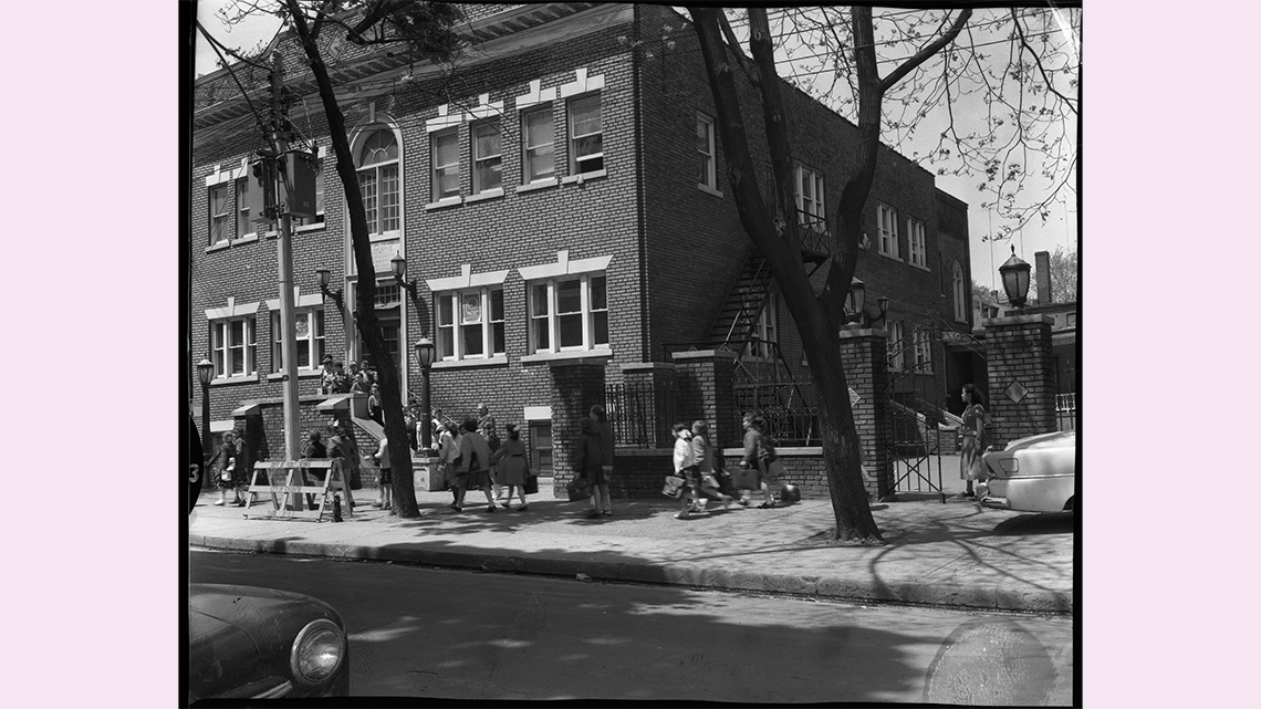A large, rectangular brick building; outside it are many schoolchildren carrying book bags.