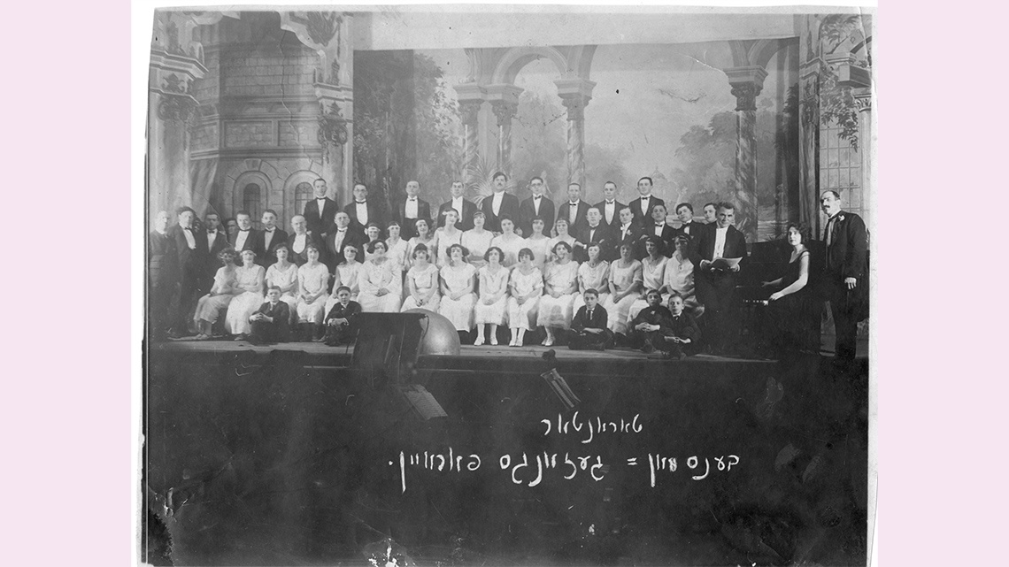 A group of women in white dresses and men in tuxedos posing for a photo in front a dramatic theatre backdrop.