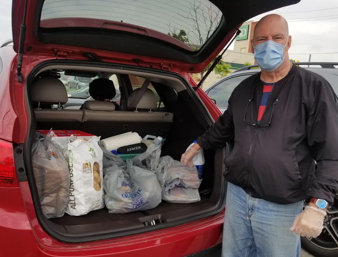 A man wearing a medical mask stands by a car loaded with groceries.